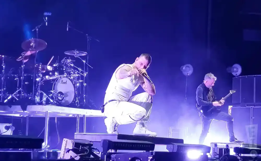 A male singer in white crouches on stage while a guitarist in black plays a guitar.