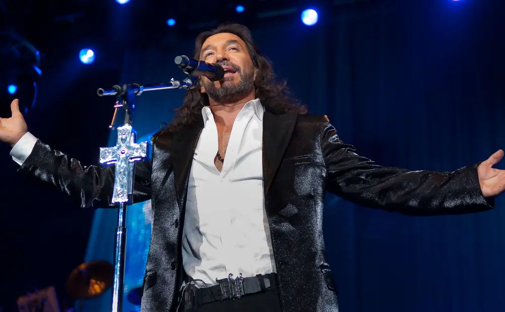 A male singer with long dark hair and a beard sings into a microphone on stage, wearing a white shirt and a sparkly black jacket.