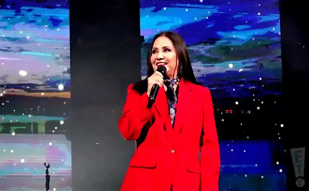 A woman in a red blazer speaks into a microphone on a stage with a starry night backdrop.