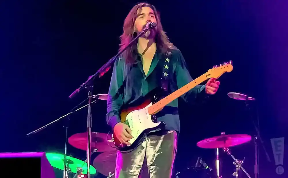 Man with long hair singing and playing a guitar on stage with colorful lighting.