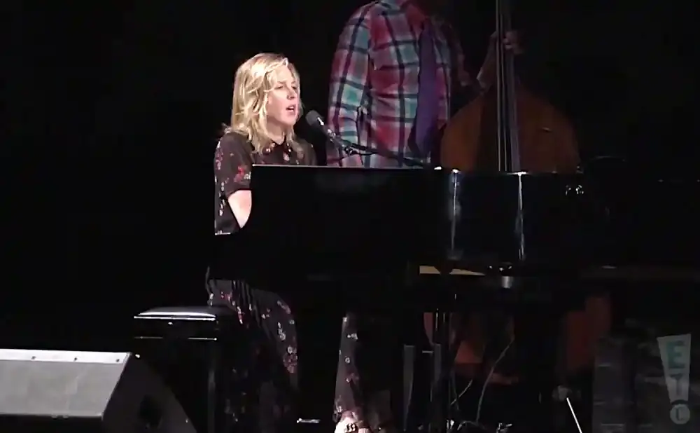 A blonde woman sings at a piano, with a man playing the double bass behind her.