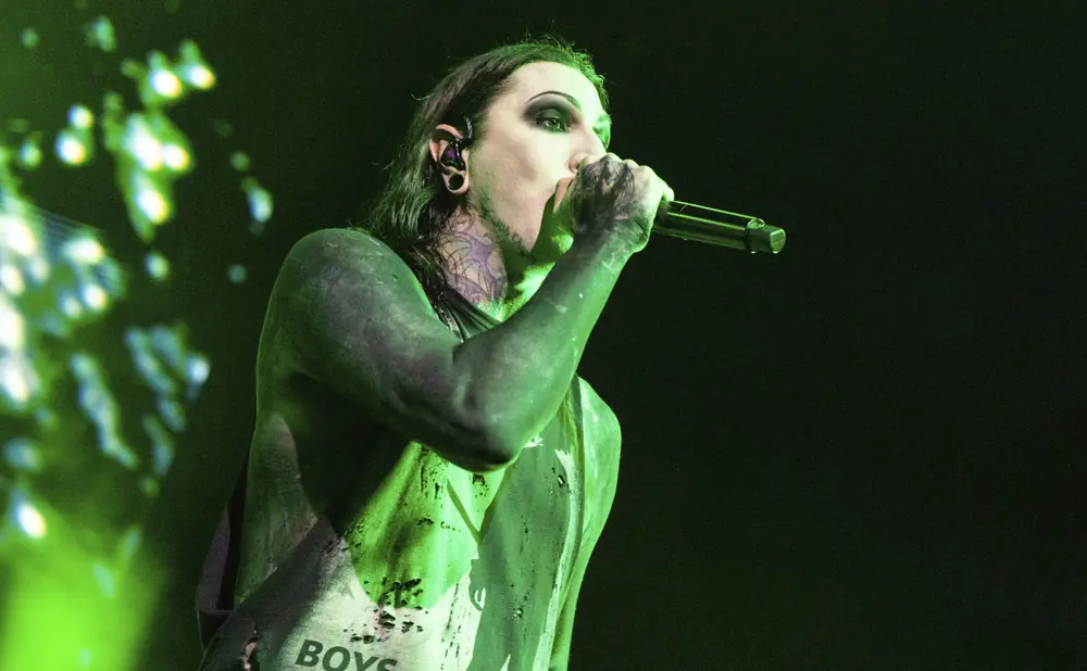 A heavily tattooed singer with green lighting and makeup sings into a microphone.