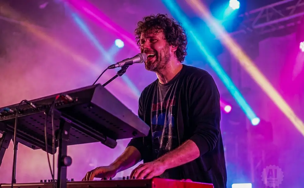 A man with curly hair sings into a microphone while playing a keyboard on a stage with colorful lights.