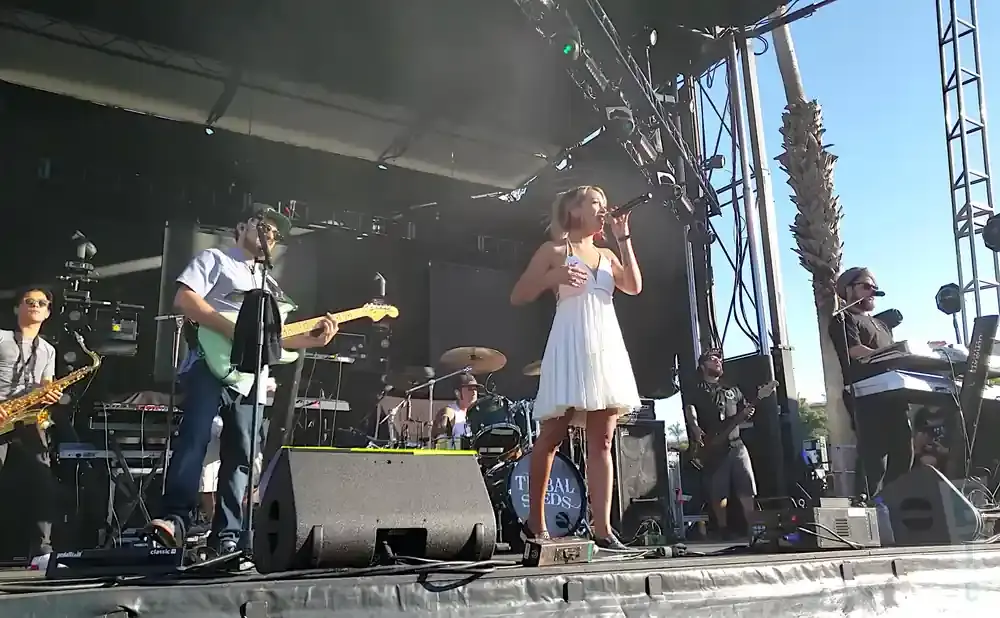 A band performs on an outdoor stage, with a female singer in a white dress, a guitarist, a saxophonist, and a keyboardist.