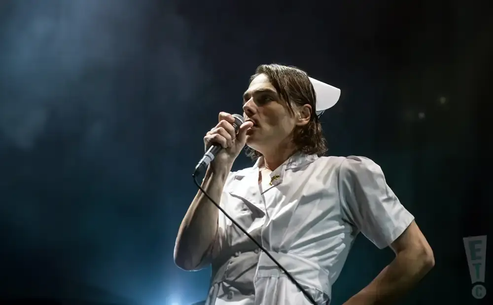 A singer in a nurse's uniform and cap performs into a microphone on a dimly lit stage.