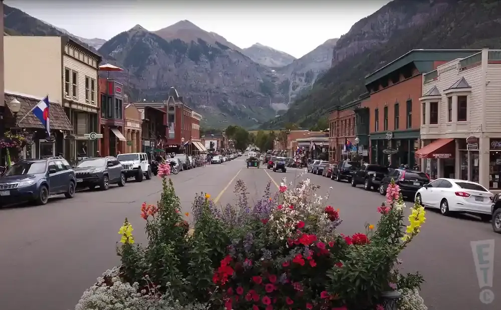 A mountain town street lined with buildings and parked cars, framed by flowers and a backdrop of majestic peaks.
