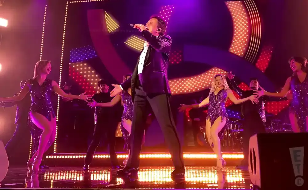 Singer in a black suit on stage with backup dancers in sequined outfits.