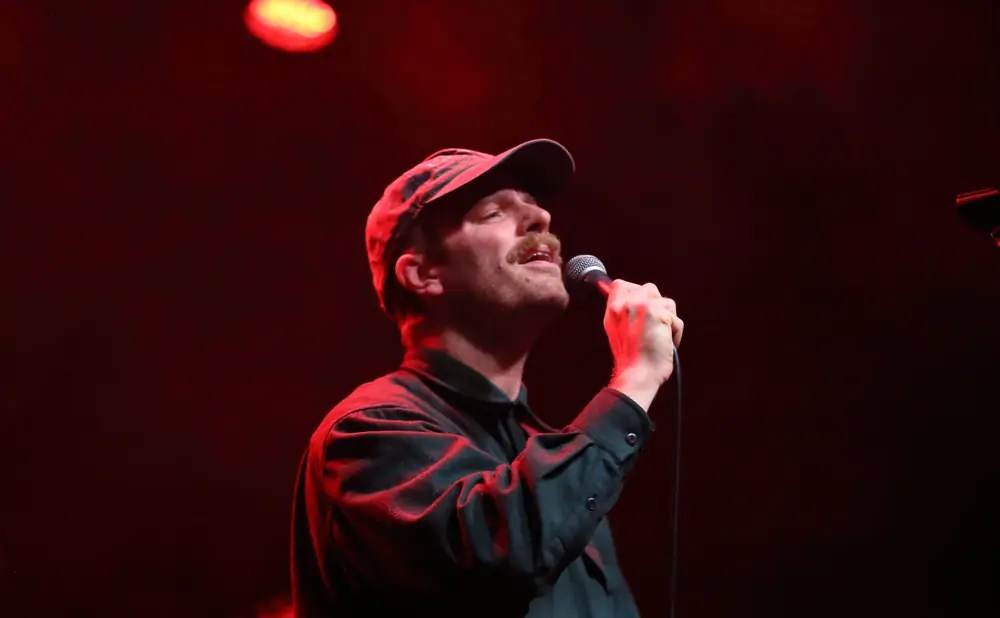 A man wearing a baseball cap and dark shirt sings into a microphone on a dimly lit stage.