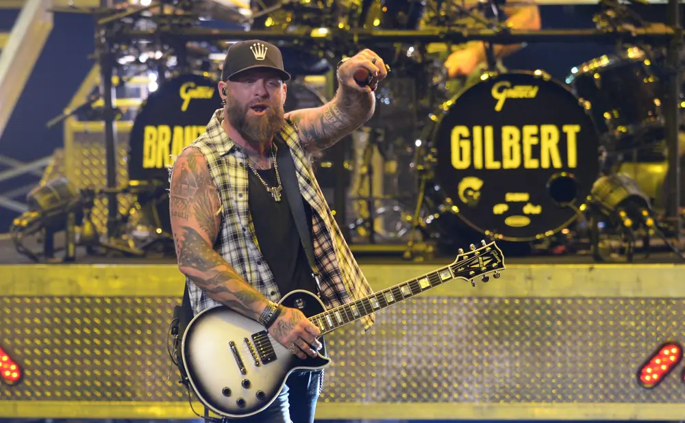 Guitarist with a beard, wearing a plaid shirt and a baseball cap, plays a white Gibson guitar on stage.