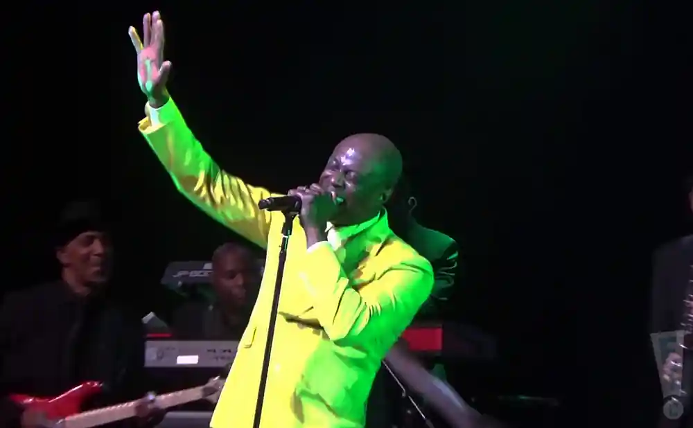 Singer in a yellow suit with his arm raised, singing into a microphone on stage.