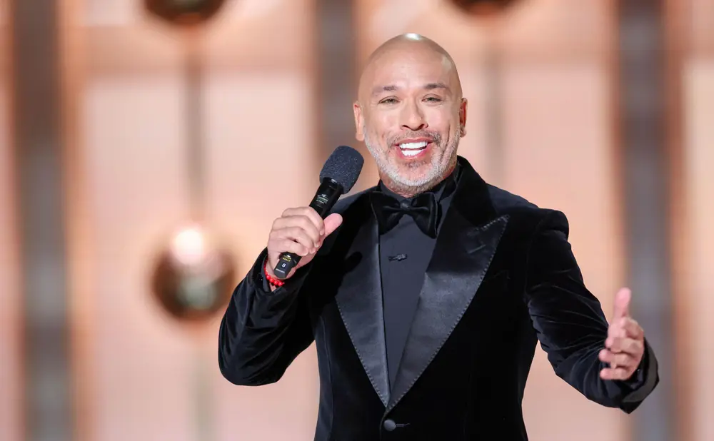 Comedian Jo Koy in a black velvet tuxedo, holding a microphone and gesturing with his hand.