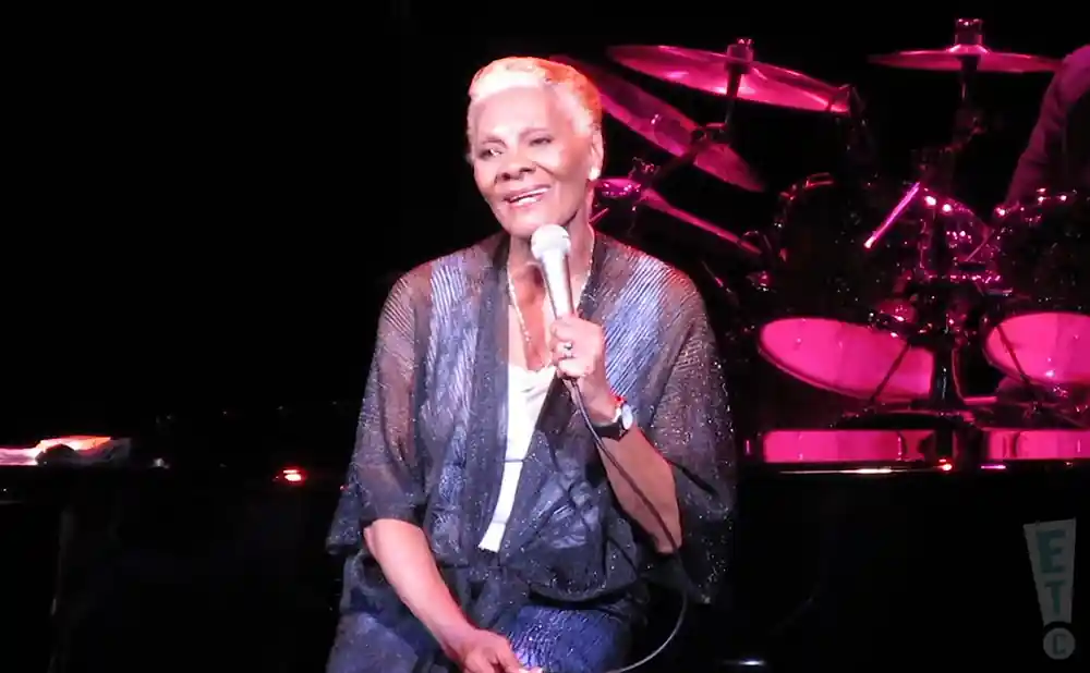 Dionne Warwick sings into a microphone on stage, wearing a shimmering robe over a white top.