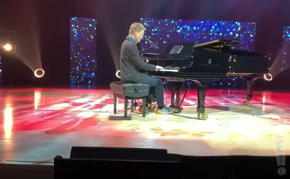 Man plays piano on stage with sparkling blue backdrop and red stage lights.
