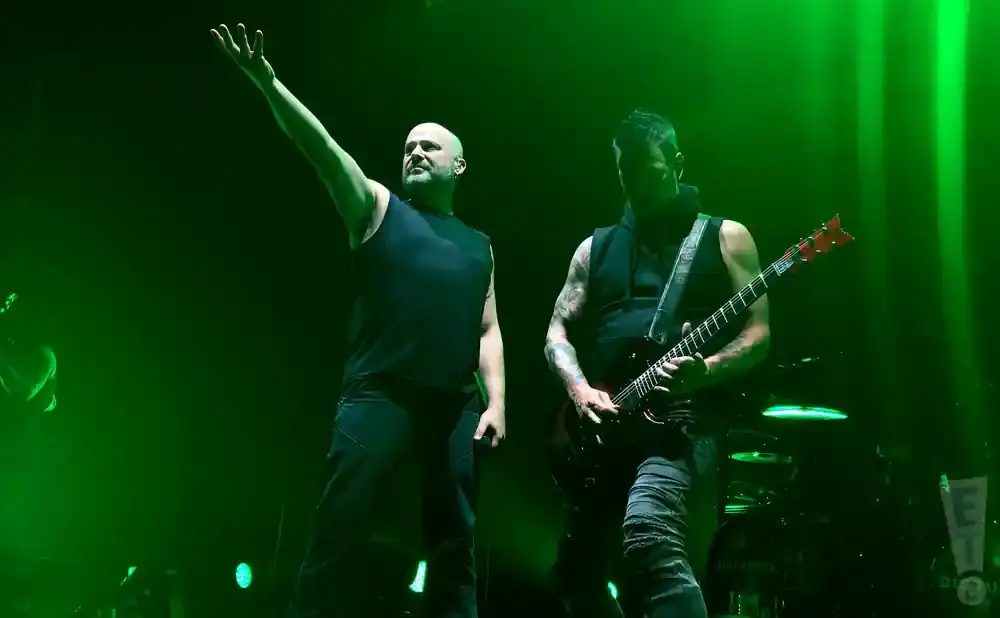 A bald singer with arm raised stands left of a guitarist playing a red electric guitar on a dimly lit stage.