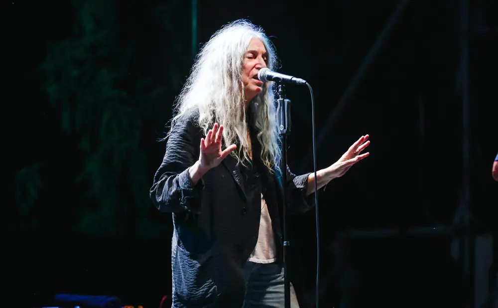 Patti Smith sings into a microphone on stage, her long white hair flowing.