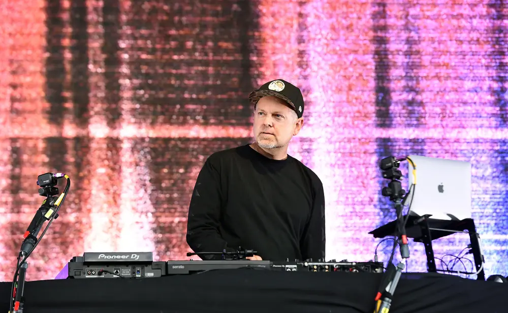DJ wearing a black t-shirt and cap, standing behind DJ equipment with a laptop and speakers.