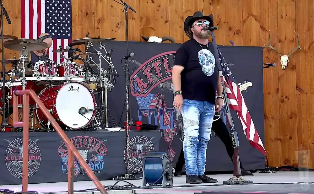 A country singer in a cowboy hat and distressed jeans performs with an American flag and skull design on his shirt.