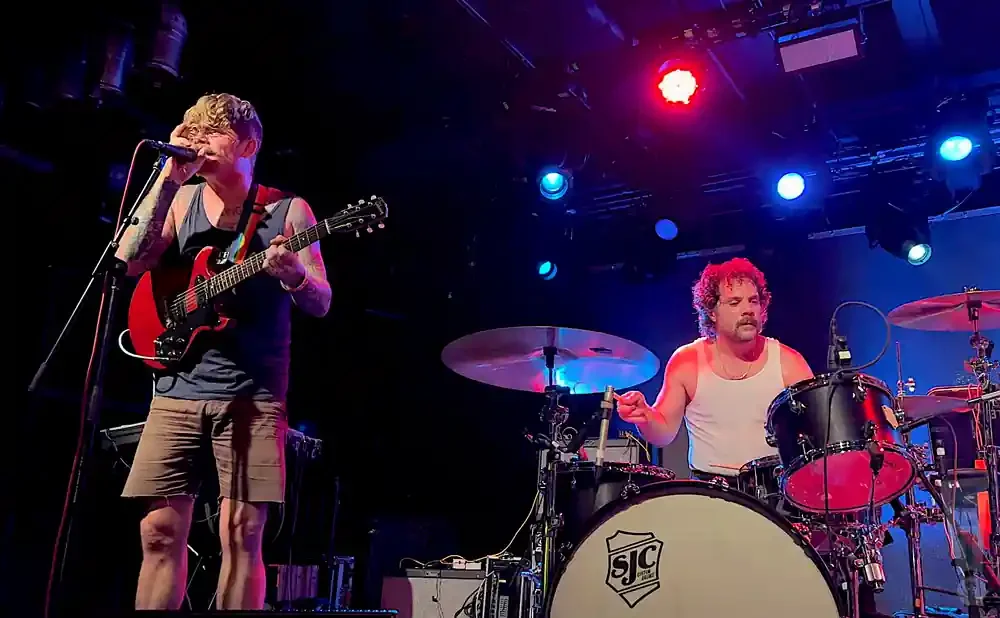 A guitarist sings into a mic while a drummer plays at a concert.