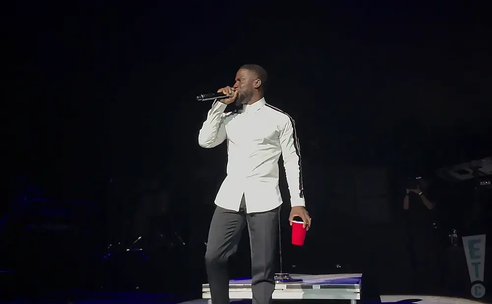 Comedian Kevin Hart on stage in a white shirt and dark pants, holding a microphone and a red cup.