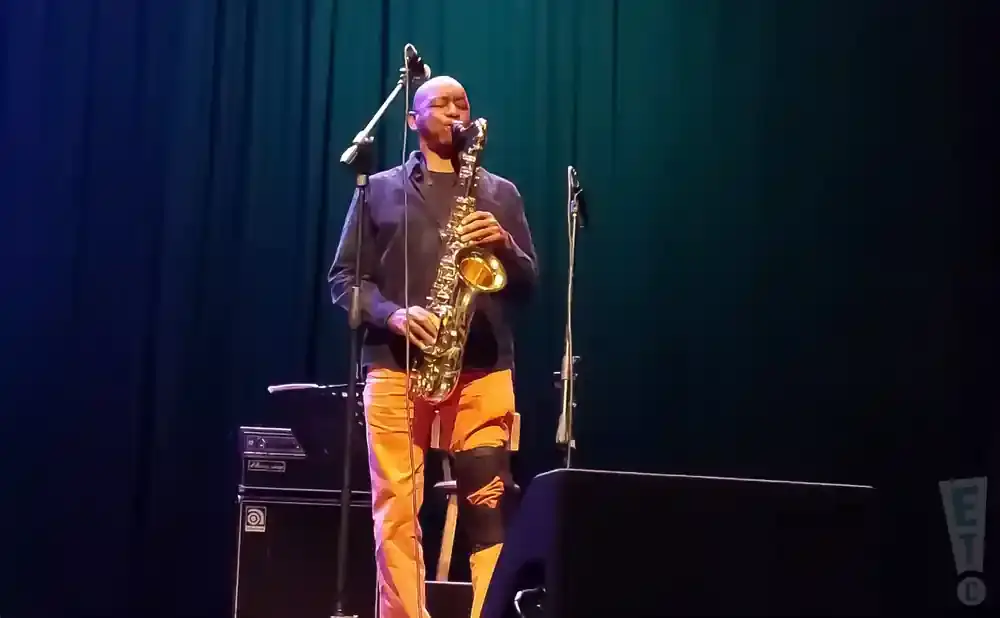 A man plays a saxophone on stage, wearing an orange outfit and a dark top.
