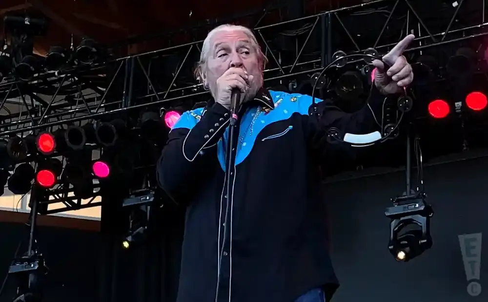 Musician with white hair wearing a black and blue shirt with a tambourine and microphone on stage.