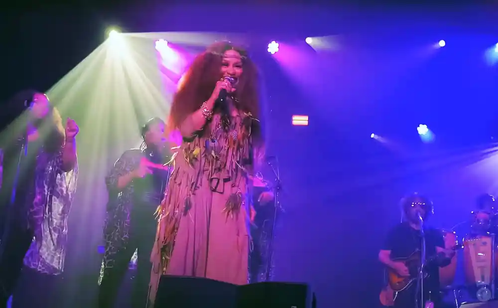 Singer with long, dark, voluminous hair sings into a microphone on a stage with a band, illuminated by green and purple lights.