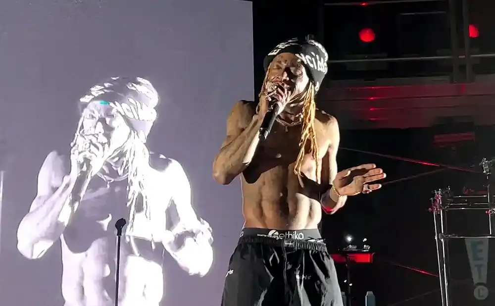 Rapper Wiz Khalifa performs on stage, shirtless, wearing a beanie and black shorts, with a large projection of himself behind him.
