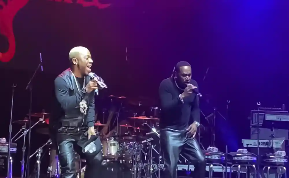 Two men in leather pants sing into microphones on a dimly lit stage with a drum set and keyboards.
