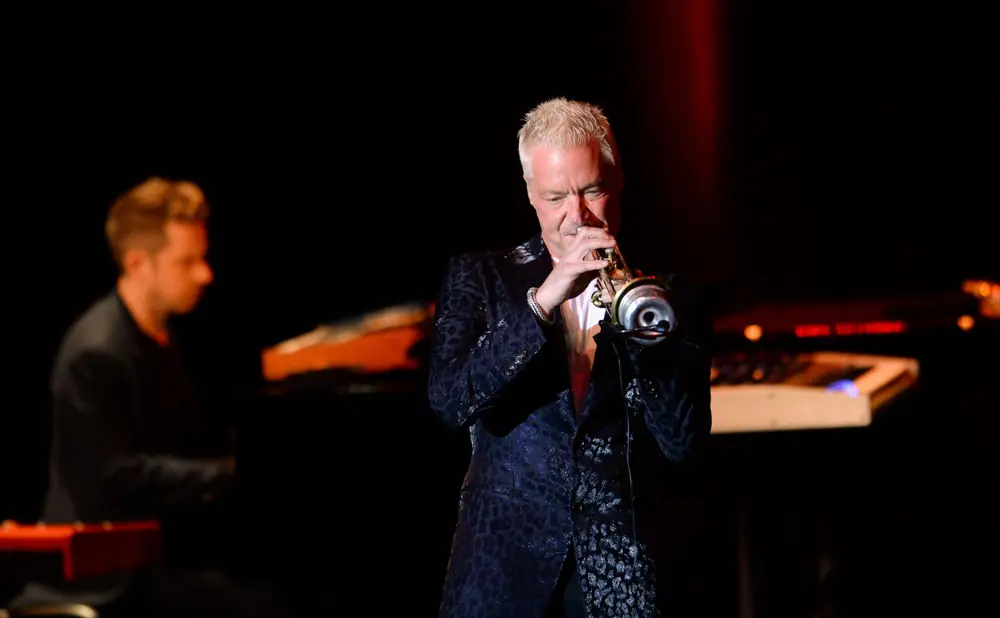 A man plays a trumpet on stage, wearing a dark jacket with a leopard-like pattern.