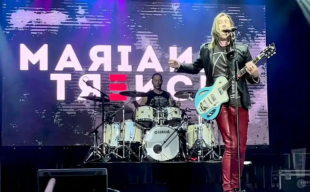 A man in a black jacket and red pants plays a blue guitar on stage with a drummer behind him.