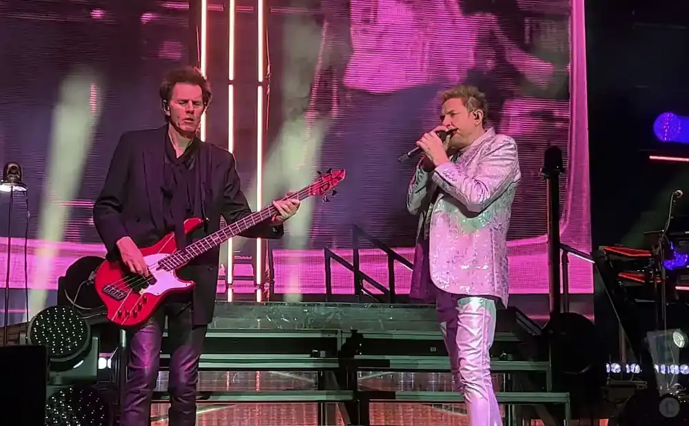Two male musicians perform on stage, one playing a red bass guitar and the other singing into a microphone.