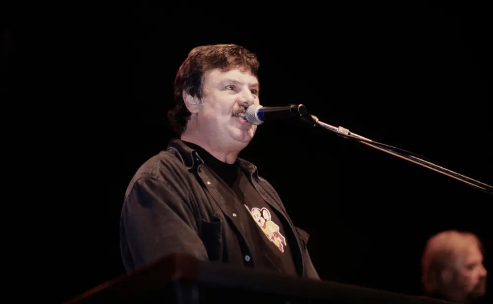 A man with a mustache sings into a microphone on stage, wearing a dark shirt and jacket.