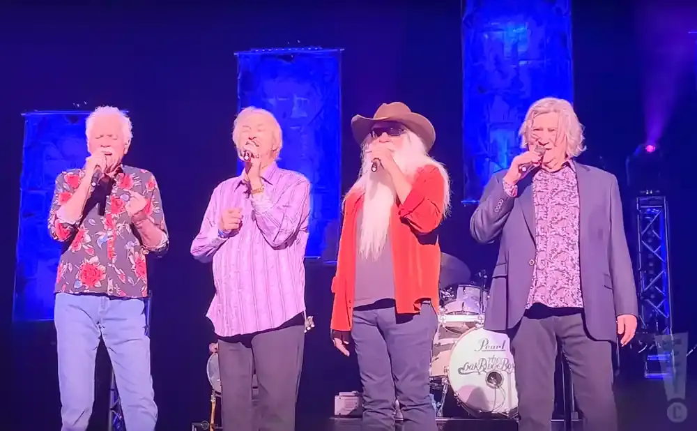 Four men singing on stage with microphones, wearing colorful shirts and jeans.