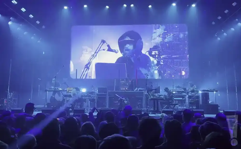 A musician performs on a stage in front of a large screen displaying their image, with a crowd watching.