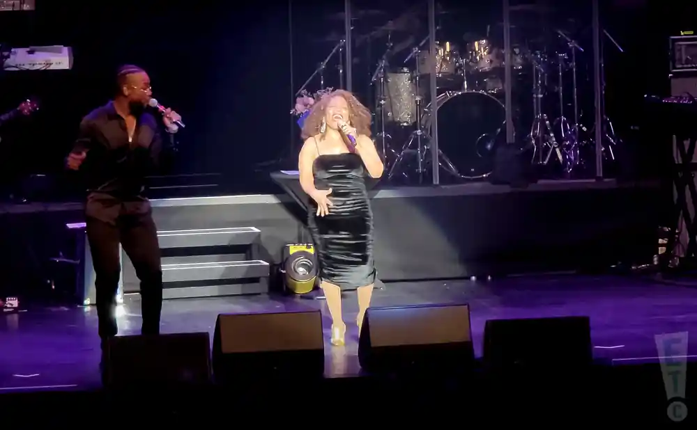 A male singer in a black shirt and pants sings into a microphone on stage next to a female singer in a black dress.