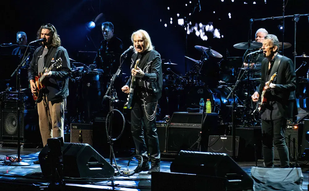 Musicians perform on stage with guitars, drums, and amplifiers, illuminated by blue stage lights.