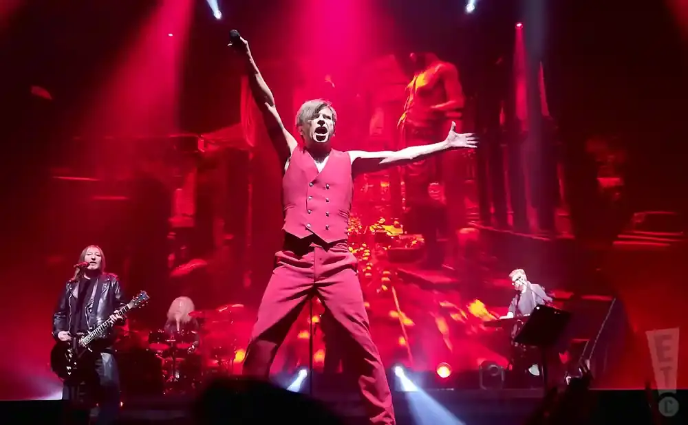 A male singer in red, arms outstretched, performs on stage with a band under red lights.