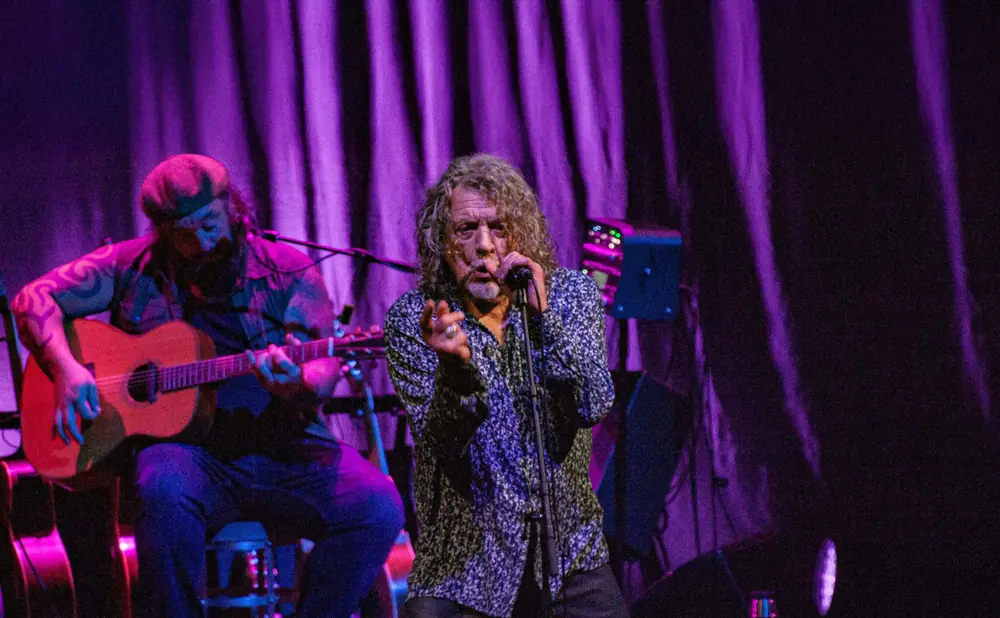 Robert Plant sings into a microphone on stage under purple lights, with a guitarist playing an acoustic guitar to his left.