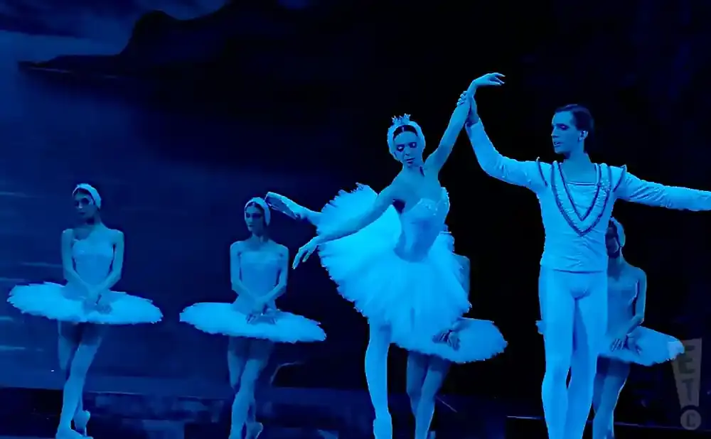 Ballerinas in white tutus and a male dancer in a light blue costume on a dark stage.