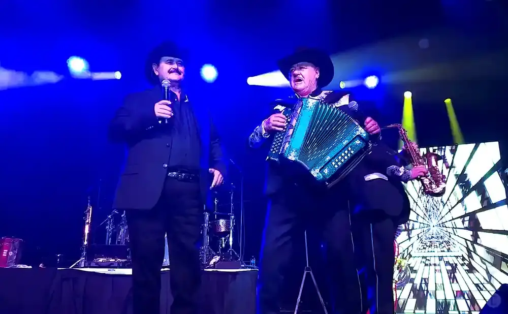Two musicians perform on stage under blue lights. The one on the left sings into a microphone, while the one on the right plays an accordion and a saxop…