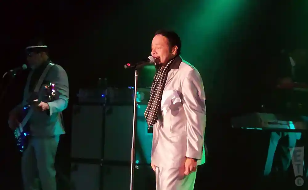 A singer in a white suit and polka dot scarf performs on stage, while a bassist in a light blue suit plays on the left.