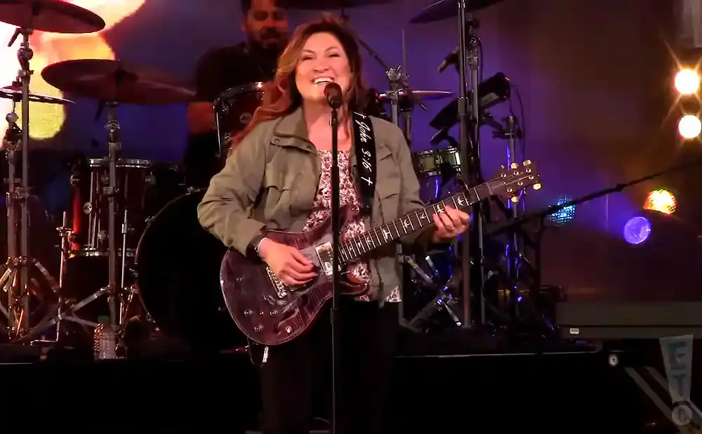 A woman with long brown hair sings and plays a purple electric guitar on stage.