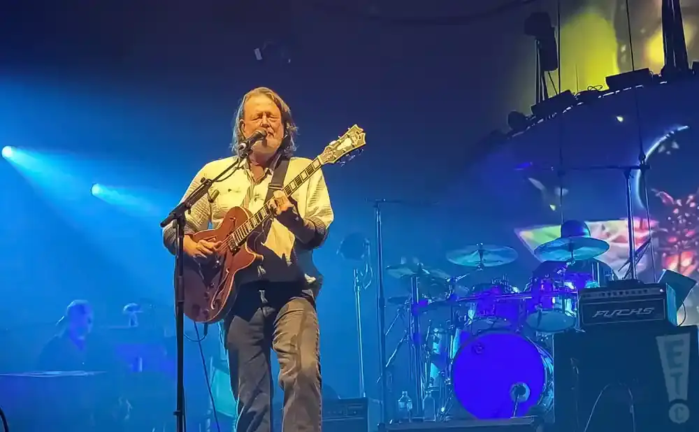 A man plays an electric guitar and sings on stage, with a drum set and keyboard player visible in the background.