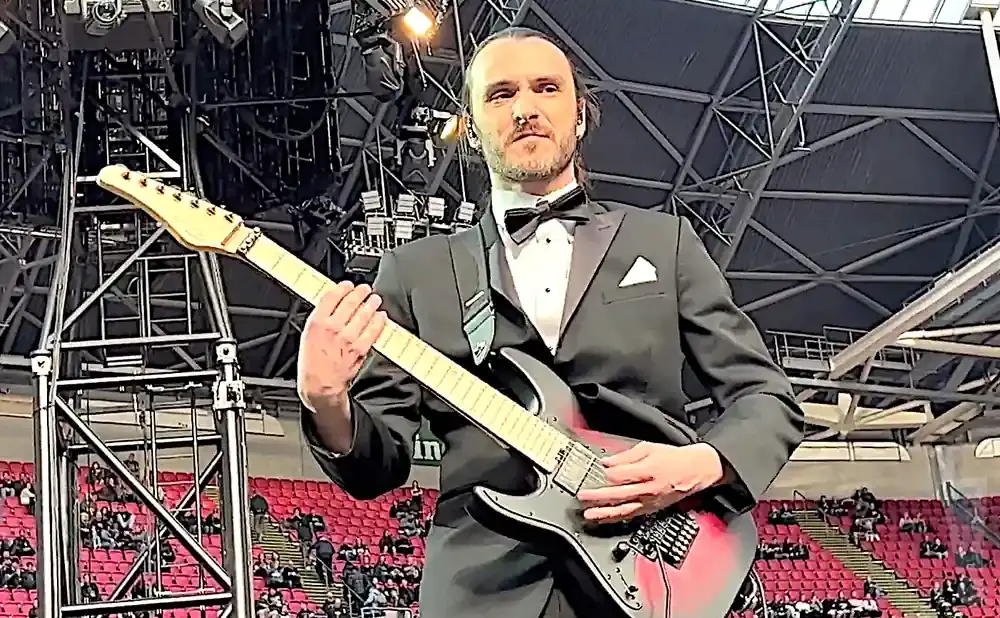 A man in a tuxedo plays a red and black electric guitar on a stage with a stadium crowd in the background.