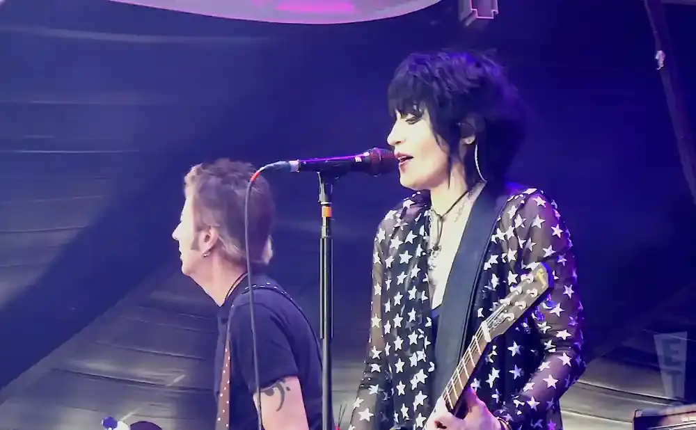 Joan Jett, in a star-print shirt, sings into a microphone while playing guitar on stage.