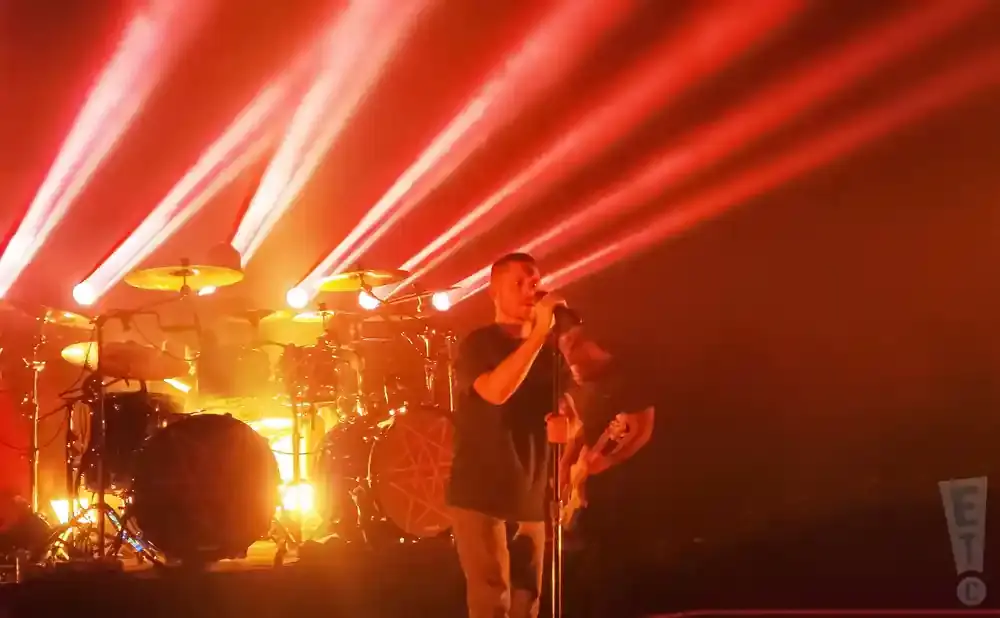 A singer on stage at a concert with red lights and a drum set behind him.