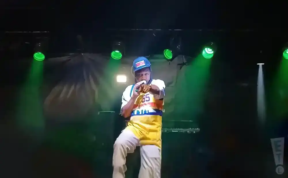 Rapper performing on stage, wearing a blue baseball cap and colorful jersey, holding a microphone.