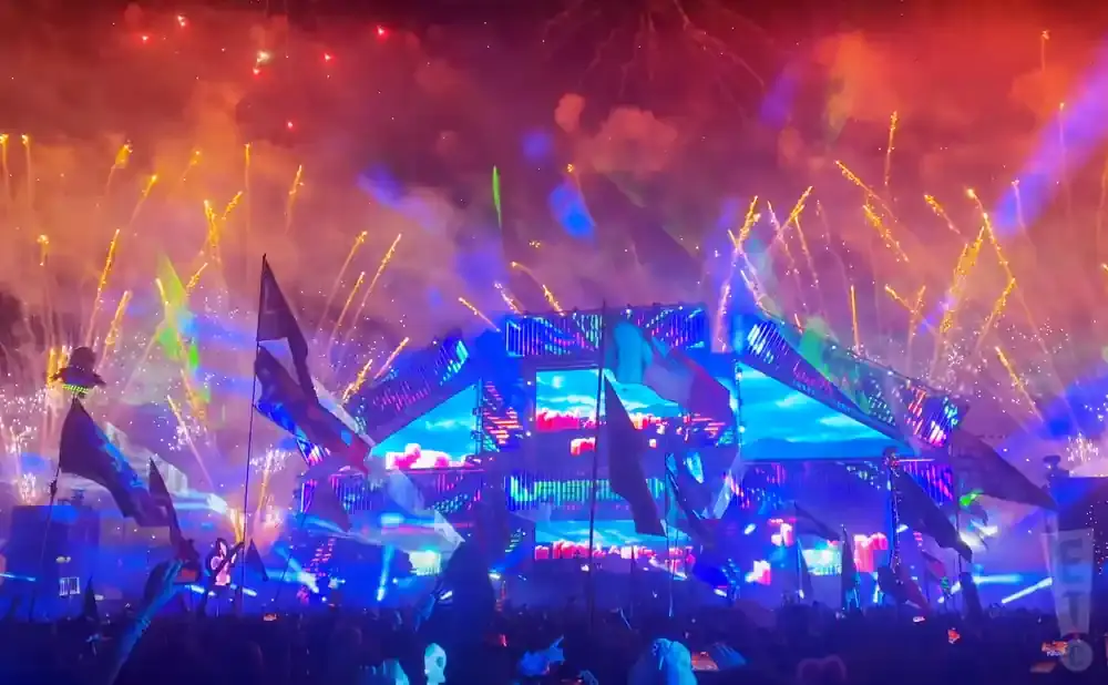 A large, bright festival stage with fireworks and flags at night.