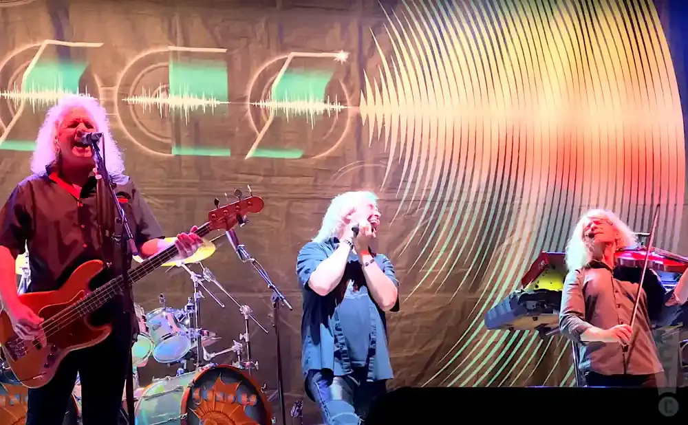 A band performs on stage, with a bassist in the foreground, a vocalist/keyboardist in the middle, and a violinist on the right.