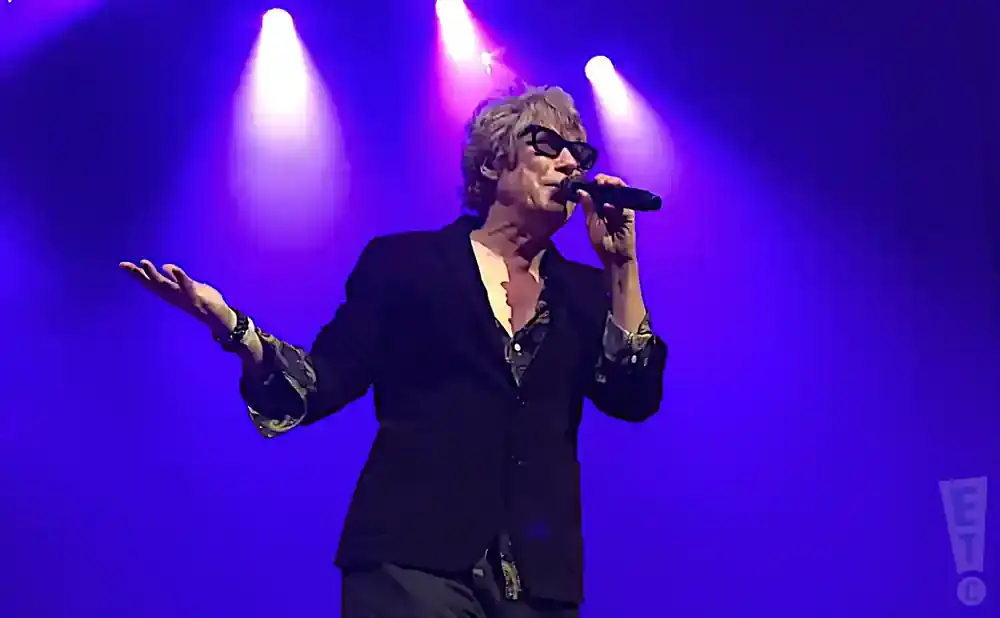 Man in black jacket and sunglasses sings into microphone on stage with purple lights.